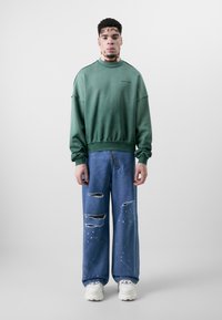 Grüner übergroßer Sweatshirt mit abgesenkten Schultern; kombiniert mit hellblauen, zerrissenen Jeans mit Farbspritzern und Rissen; weiße Sneakers.
