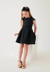 Schwarzes Kleid mit kurzen Ärmeln und weitem Rock, strukturierter Taillenbund, kombiniert mit goldenen T-Riemenschuhen und einem passenden schwarzen Schleifenhaarband.