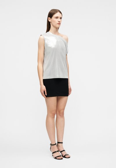 Νεαρή γυναίκα στέκεται φορώντας αμάνικο ασημί τοπ με έναν ώμο ακάλυπτο, μαύρη μίνι φούστα και μαύρα σανδάλια με λουράκια.