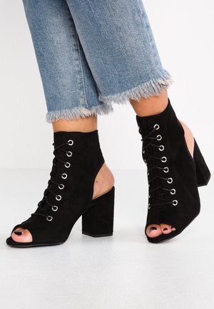 Bottes à talons hauts en daim noir à bout ouvert avec lacets, portées avec un jean bleu clair raccourci à ourlet effiloché sur une surface blanche.