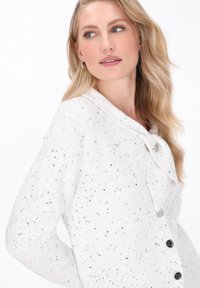 White knit cardigan with black speckles. Features a shawl collar, front buttons, and a soft texture. Classic fit with a relaxed design.