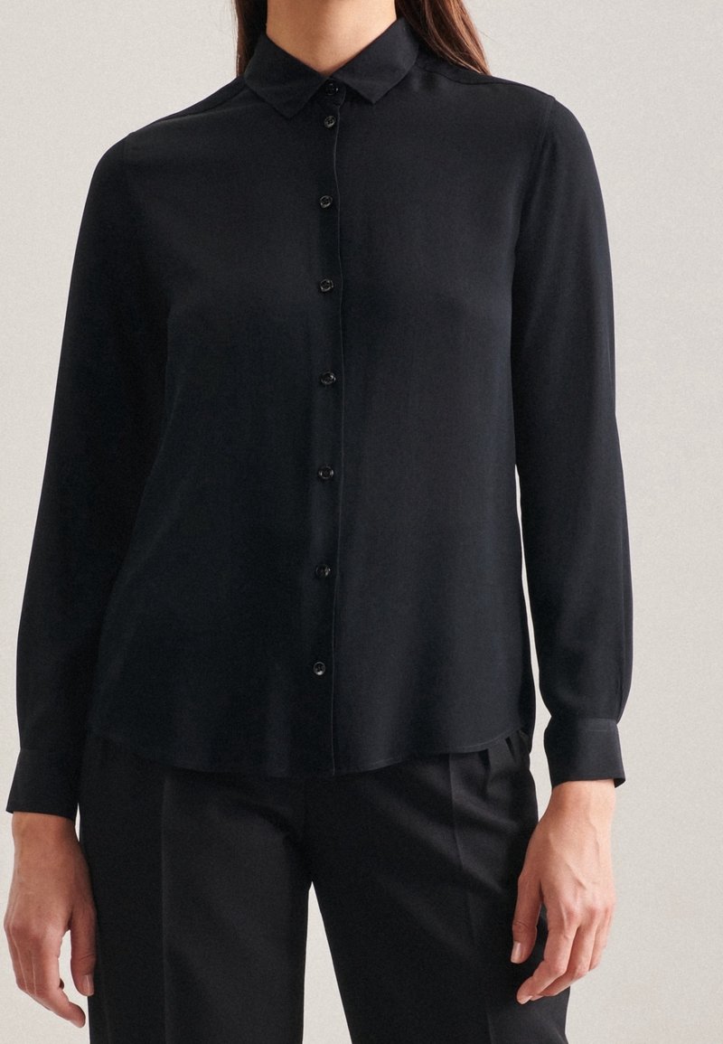 Femme portant une chemise noire à manches longues avec col et boutons, ainsi qu'un pantalon noir, debout devant un fond clair uni.