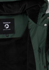 Binnenkant van een groen en zwart jack met een label "Travelin' where we walk" en een maat medium, dat de rits en drukknoopsluitingen toont.