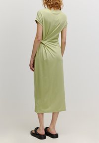 Knie-langes, limonengrünes Kleid aus weichem Stoff. Mit einer Drehung am Rücken, kurzen Ärmeln und einem lockeren Schnitt. Getragen mit schwarzen Sandalen.