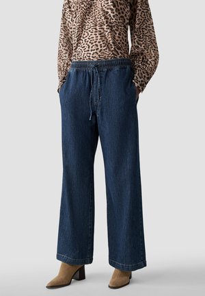 Person trägt dunkelblaue Jeans mit weitem Bein, elastischem Bund und Kordelzug, kombiniert mit hellbraunen Stiefeletten und einem Oberteil mit Leopardenmuster.