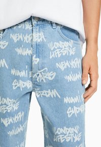 Ljusblå denimjeans med ett vitt graffiti-inspirerat textmönster med stadnamn. Klassisk femfickdesign med metallknappstängning.