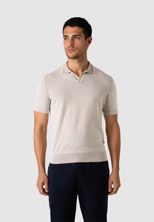 Mann, der ein hellbeiges Kurzarm-Poloshirt und eine dunkle Hose trägt, vor einem schlichten hellgrauen Hintergrund steht und zur Seite schaut.