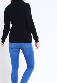 Černý pletený svetr s vysokým límcem, dlouhými rukávy a strukturovaným vzorem, kombinovaný s těsně přiléhajícími modrými džíny.