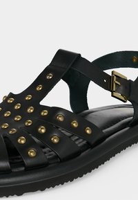 Μαύρο δερμάτινο σανδάλι με χρυσά στρογγυλά καρφιά, πολλαπλούς ιμάντες, ανατομική σόλα και ρυθμιζόμενο χρυσό κούμπωμα.