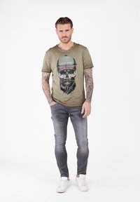 Camiseta verde oliva con un gráfico de calavera y diseño de gorra, combinada con jeans ajustados y desgastados de color gris y zapatillas blancas.