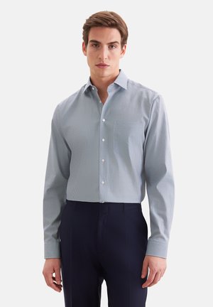 Mann trägt ein hellblau gemustertes, langärmeliges Hemd und dunkelblaue Hose vor einem einfarbigen Hintergrund.