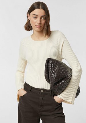 Młoda kobieta o krótkich brązowych włosach ma na sobie kremowy prążkowany sweter i ciemne spodnie, trzymając pod pachą dużą czarną plecioną kopertówkę.