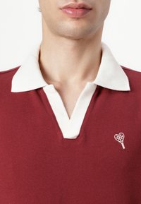 Kruvino polo marškiniai su baltu apvadu, prie krūtinės turi mažą siuvinėtą širdelės dizainą. Pagaminti iš tekstūruoto medvilnės audinio.