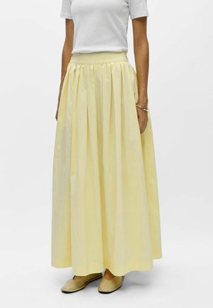 Femme portant une longue jupe plissée jaune pâle, un haut blanc à manches courtes, des chaussures plates beiges et un bracelet au poignet droit.