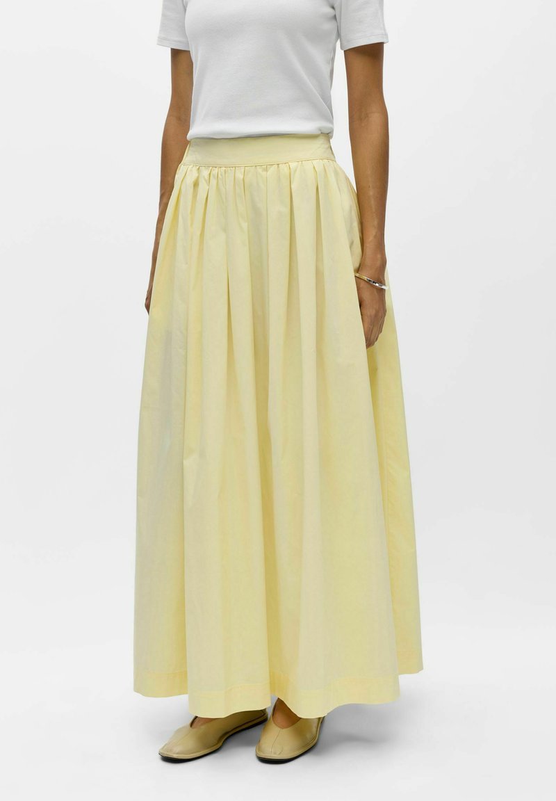 Femme portant une longue jupe plissée jaune pâle, un haut blanc à manches courtes, des chaussures plates beiges et un bracelet au poignet droit.