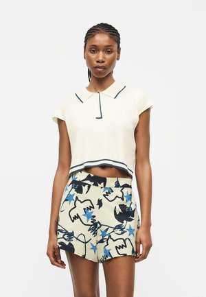 Γυναίκα με πλεγμένα μαλλιά που φορεί ένα κοντό λευκό πόλο μπλουζάκι και μπεζ σορτς με μπλε αστέρι και μαύρα αφηρημένα σχέδια πουλιών.