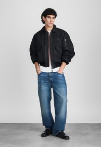 Black bomber jacket with zip pockets, worn over a white T-shirt, paired with loose blue jeans and black shoes. Smooth fabric with a relaxed fit.