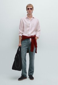 Lichtroze linnen shirt, rode trui om de taille geknoopt, blauwe denim jeans, zwarte stoffen tas, donkere schoenen, vierkante zonnebril.