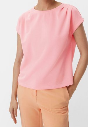 Femme portant un chemisier rose à manches courtes associé à un pantalon taille haute couleur pêche, une main dans la poche sur un fond uni