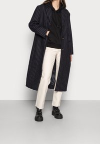 Abrigo largo a rayas azul marino con dos botones, superpuesto a una sudadera negra, combinado con pantalones cortos blancos y botas negras gruesas.