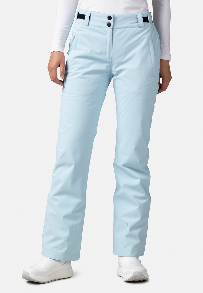 Ljust blå skidbyxor med en slät textur, två framfickor och två knappstängningar i midjan. Åtföljs av vita skor.