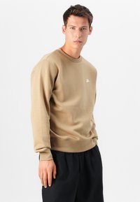 Bruine sweatshirts met ronde hals, ribboord bij de manchetten en onderkant. Witte Nike-logo op de borst. Draagt met zwarte shorts. Gladde textuur.