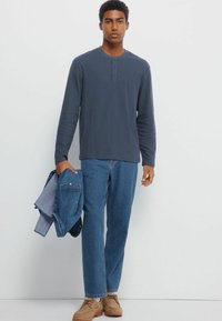 Langärmliges navy-blaues Henley-Shirt mit drei Knöpfen, kombiniert mit hellblauen Jeans und braunen Wildlederschuhen, hält eine Jeansjacke.