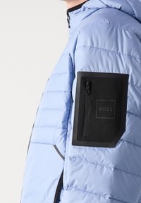 Ljusblå puffjacka med horisontell sömnad, med en svart dragkedjeficka på vänster arm och en minimalistisk logotyp på fickan.