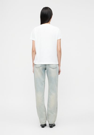 Άτομο με μακριά μαύρα μαλλιά που φοράει ένα απλό λευκό κοντομάνικο μπλουζάκι και ανοιχτό μπλε τζιν παντελόνι με ίσια μπατζάκια, στέκεται γυρισμένο προς τα πίσω.