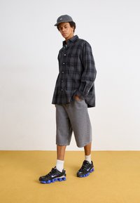 Fekete és szürke kockás flaneling, szürke sort, fekete sneaker kék részletekkel, fehér zoknival. Egyszínű háttér.