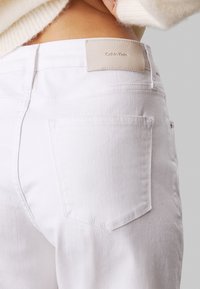 Białe dżinsy o gładkiej teksturze, z średnim stanem i skórzaną metką z napisem "Calvin Klein" z tyłu. Widoczne dwie tylne kieszenie.