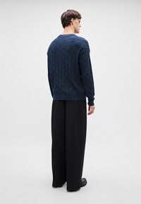 Mørkeblå teksturert genser med geometrisk mønster, ribbede mansjetter og kant, kombinert med brede sorte bukser og svarte sko.