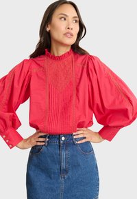 Blusa roja con un cuello con volantes, intrincados bordados y mangas abullonadas. Combinada con una falda de mezclilla, que muestra un estilo casual.