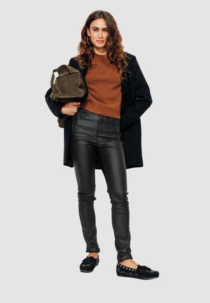 Pantalons en simili cuir noir, haut en tricot de couleur rouille, manteau noir oversize et ballerines noires avec détails métalliques, accompagnés d'un sac vert olive.