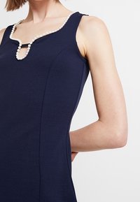 Granatowa sukienka bez rękawów z dekoltem wykończonym perełkami oraz detalem w kształcie dziurki na piersi. Gładki, dopasowany materiał z bocznymi szwami.