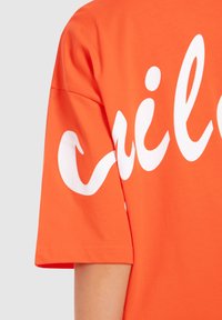Oranžové bavlněné tričko s velkým bílým psaným textem, krátkými rukávy a uvolněným střihem. Lehká textilie, jednoduché švy.