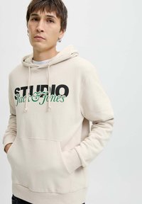 Svijetložuti hoodie s crno-zelenim grafičkim dizajnom koji glasi "STUDIO Jack & Jones." Sadrži džep za kengure i kapuljaču s vezicom.