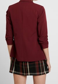 Burgundy blazer with three-quarter sleeves, tailored fit, and a smooth finish, paired with a black and red plaid skirt featuring a fringe hem.