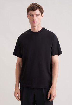 Junger Mann mit kurzen braunen Haaren trägt ein schlichtes schwarzes T-Shirt mit rundem Ausschnitt und kurzen Ärmeln sowie schwarze Hosen und steht vor einem schlichten hellen Hintergrund.