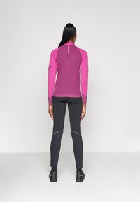 Getailleerd overhemd met lange mouwen in roze en paars, vochtafvoerende stof, platte naden, met logo op de rug. Gecombineerd met donkere leggings.