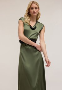 Abito in satin verde oliva con maniche a cappuccio, dotato di una cintura nera e una collana massiccia nera e oro. Tessuto liscio, design lungo fino al pavimento.