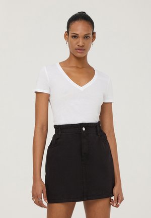 T-shirt bianco a V abbinato a una mini gonna in denim nera. La gonna presenta un alta vita, tasche laterali e chiusura con bottone.