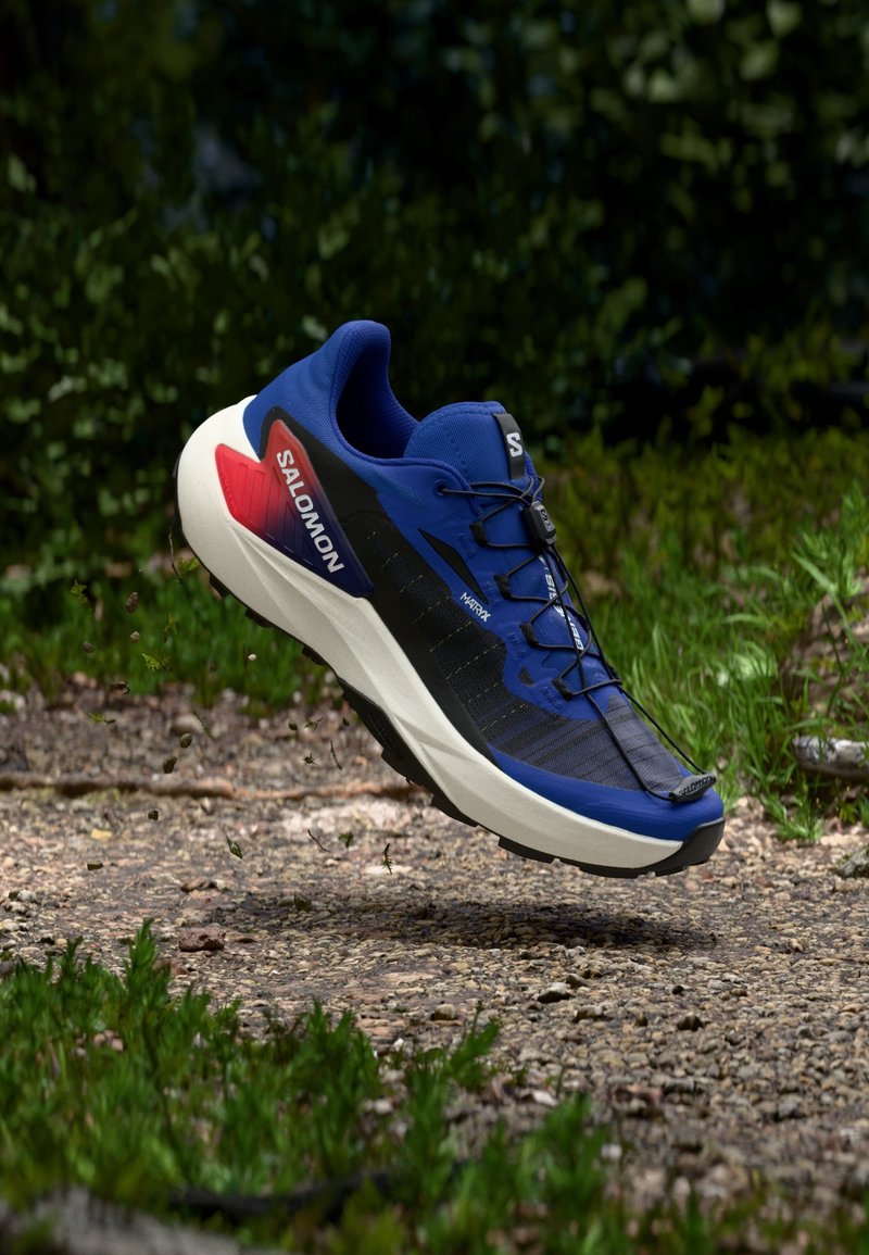 Chaussure de trail running Salomon bleue, noire et rouge flottant au-dessus d'un chemin de terre avec de la verdure en arrière-plan.