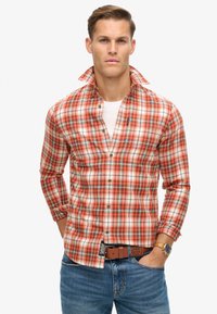 Rot- und orangefarbenes Karohemd mit langen Ärmeln, Button-Down-Kragen und Brusttasche. Über einem weißen T-Shirt getragen, kombiniert mit blauen Jeans.