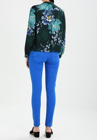 Blouse florale verte avec des accents bleus, et jean skinny bleu vif. L'ensemble se compose de manches longues et d'une coupe ajustée, associé à des talons noirs.