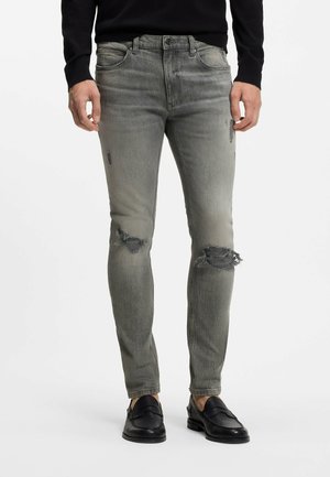 Mann trägt graue Slim-Fit-Jeans mit Rissen an den Knien, schwarzen Pullover und schwarze Leder-Loafer, steht vor weißem Hintergrund.