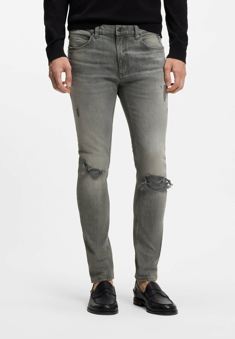 Homme portant un jean gris slim déchiré aux genoux, un pull noir et des mocassins en cuir noirs, debout sur un fond blanc.