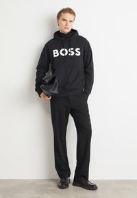 Jonge man in zwarte hoodie met tekst "BOSS", zwarte broek en schoenen, houd een zwarte gevouwen leren tas vast, staand tegen een witte muur.