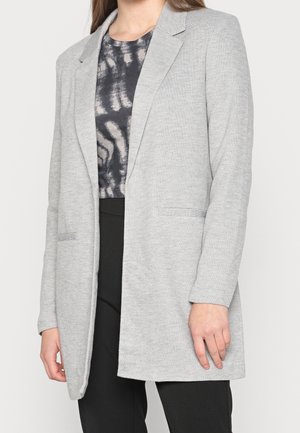 Grijze blazer met geknipte revers, open voorkant en twee zijzakken. Gemaakt van zachte stof met een gladde textuur, gecombineerd met een donker patroon top.