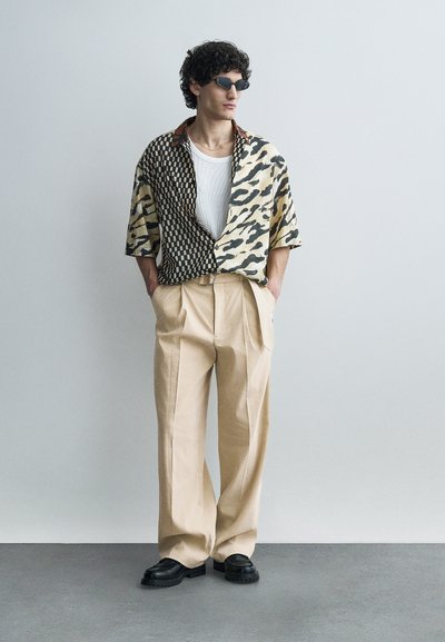 Άνδρας με σγουρά μαλλιά και γυαλιά ηλίου, ντυμένος με πουκάμισο κοντομάνικο με σχέδια πάνω από λευκό τοπ, μπεζ πλισέ παντελόνι και μαύρα παπούτσια, στέκεται μπροστά σε απλή τοίχο.